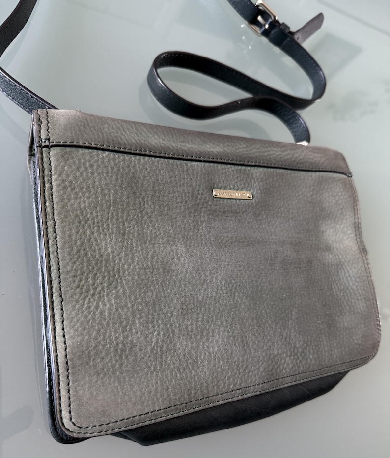 May include: A rectangular crossbody bag featuring a textured grey leather flap and a smooth black leather base. The bag includes a thin black adjustable shoulder strap with a silver-toned metal buckle and a small rectangular silver logo plate.