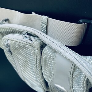 K&ouml;nnte beinhalten: Eine silberne und graue Bauchtasche mit einem Rei&szlig;verschlussfach und einem schwarzen verstellbaren G&uuml;rtel mit einer Schnalle.