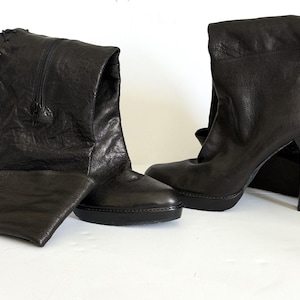May include: A pair of black leather boots. One boot has a folded-over cuff and a zipper, while the other has a high heel and lace-up detailing on the back. The boots are on a white surface.