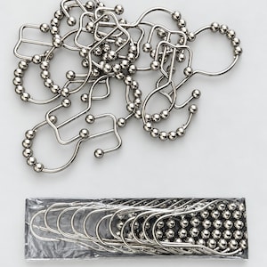 May include: A collection of silver-toned metal shower curtain hooks, each featuring a series of small decorative spheres along the curved frame. Some hooks are loose and scattered, while others are neatly stacked inside a clear plastic sleeve.
