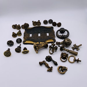 May include: A collection of antique brass cabinet knobs, pulls, and a light switch plate. The knobs and pulls feature ornate designs, including floral, scroll, and lion head motifs. The switch plate is black with gold accents.