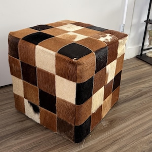 May include: A cube-shaped ottoman with a patchwork design. The ottoman is covered in squares of cowhide in shades of brown, black, and cream. The patchwork pattern creates a geometric design. This decorative piece can be used as a footrest or extra seating.