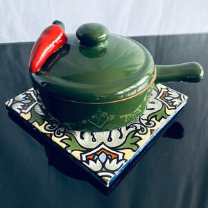 May include: A green ceramic casserole dish with a red chili pepper lid. The dish is sitting on a colorful tile trivet.