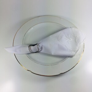 May include: A white napkin with an embroidered design is placed on a white plate with a gold rim.