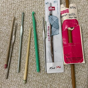 May include: A set of six knitting needles and crochet hooks, including a wooden cable stitch holder. The needles are made of metal and wood, and the crochet hooks are made of plastic. The cable stitch holder is made of wood and has a red and white label that says "Scovill Hero Cable Stitch Holder 65¢".