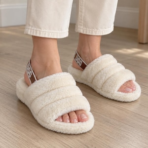 May include: Cream-colored fluffy slippers featuring a wide, padded strap across the top and a brown elastic heel strap with the white text UGG printed on it. The slippers have a thick, plush texture and an open-toe design.