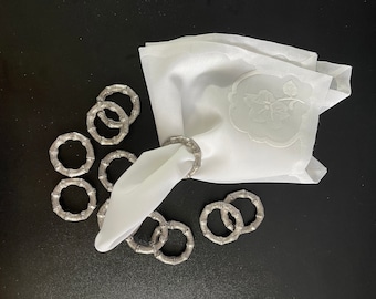 May include: A set of silver napkin rings with a bamboo design. The rings are arranged on a black surface with a white napkin with an embroidered floral design.