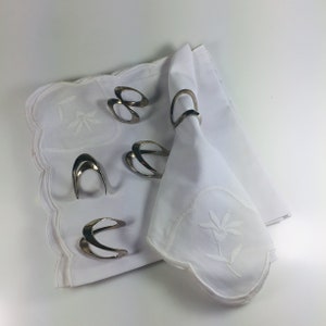 May include: Five silver napkin rings on a white tablecloth with an embroidered floral design. The napkin rings are made of a smooth, shiny metal and have a simple, elegant design.