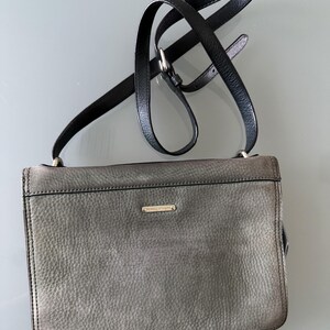 May include: A rectangular crossbody bag in a textured grey leather finish with a black leather adjustable shoulder strap. The bag features a small, horizontal gold-toned metal plaque centered on the front flap and black leather trim along the edges.