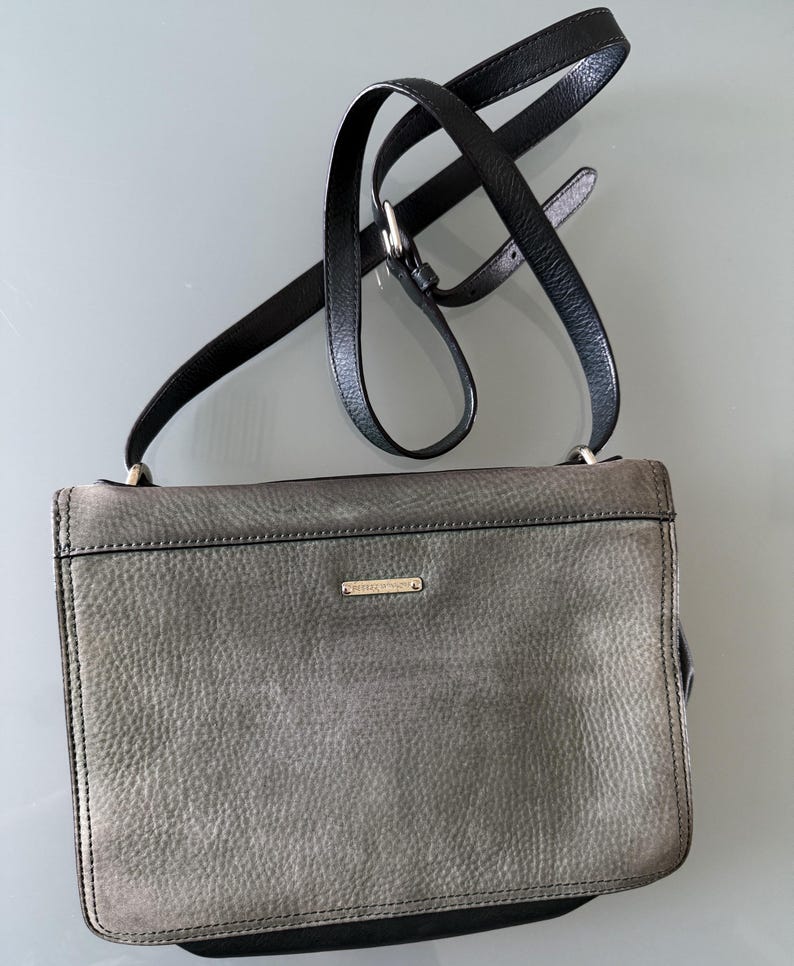 May include: A rectangular crossbody bag in a textured grey leather finish with a black leather adjustable shoulder strap. The bag features a small, horizontal gold-toned metal plaque centered on the front flap and black leather trim along the edges.