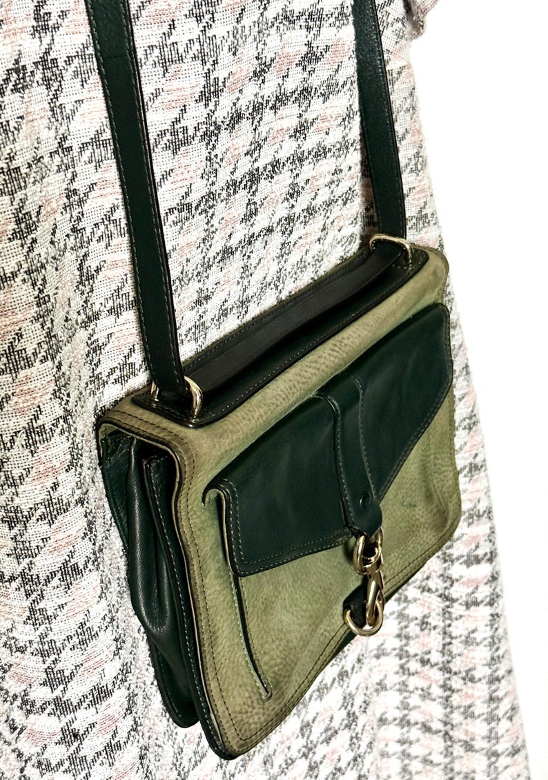May include: A crossbody bag featuring a two-tone design with dark forest green leather accents and a lighter sage green textured leather body. The bag includes a dark green shoulder strap, a front flap pocket, and a gold-toned metal swivel clasp.