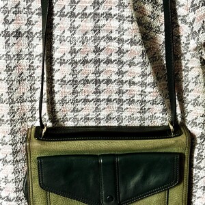 May include: A rectangular olive green leather crossbody bag with a black leather flap pocket on the front. The bag features a gold-toned metal lobster clasp closure and a long black leather shoulder strap, showcasing a textured leather finish.