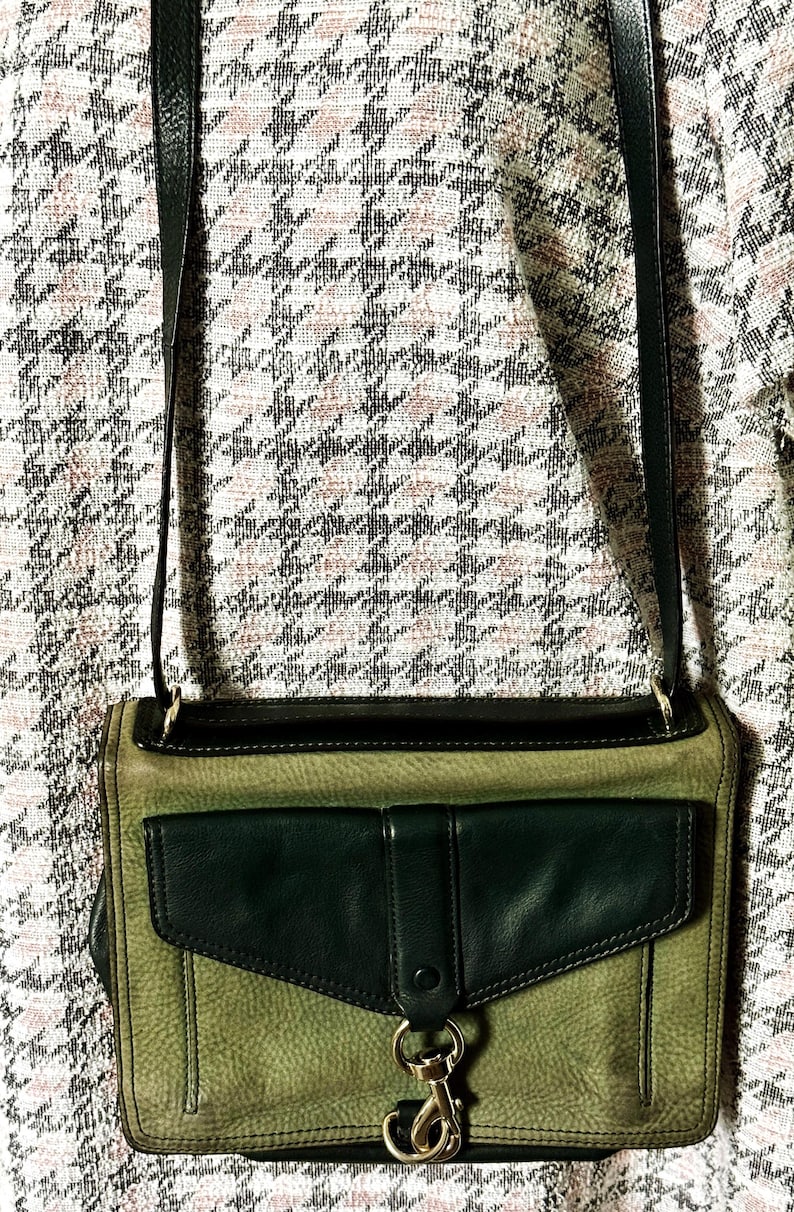 May include: A rectangular olive green leather crossbody bag with a black leather flap pocket on the front. The bag features a gold-toned metal lobster clasp closure and a long black leather shoulder strap, showcasing a textured leather finish.