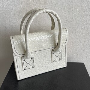 May include: A white faux leather handbag with a crocodile embossed pattern. The bag has two top handles and a flap closure with a decorative X stitch detail.