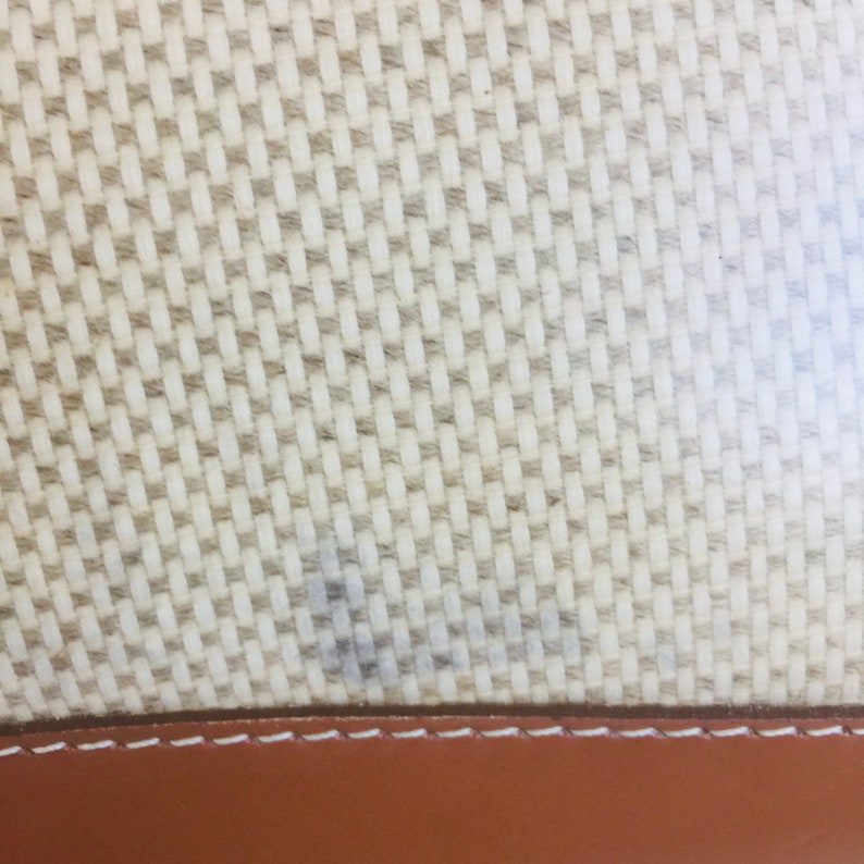 May include: Close-up of a beige woven fabric with a brown leather trim. The fabric has a subtle pattern of small squares.