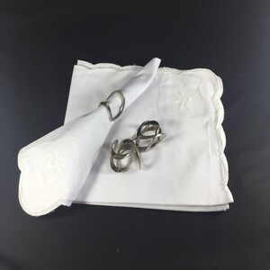 May include: Two silver napkin rings on a white linen napkin with embroidered floral designs. The napkin is folded into a triangle shape.