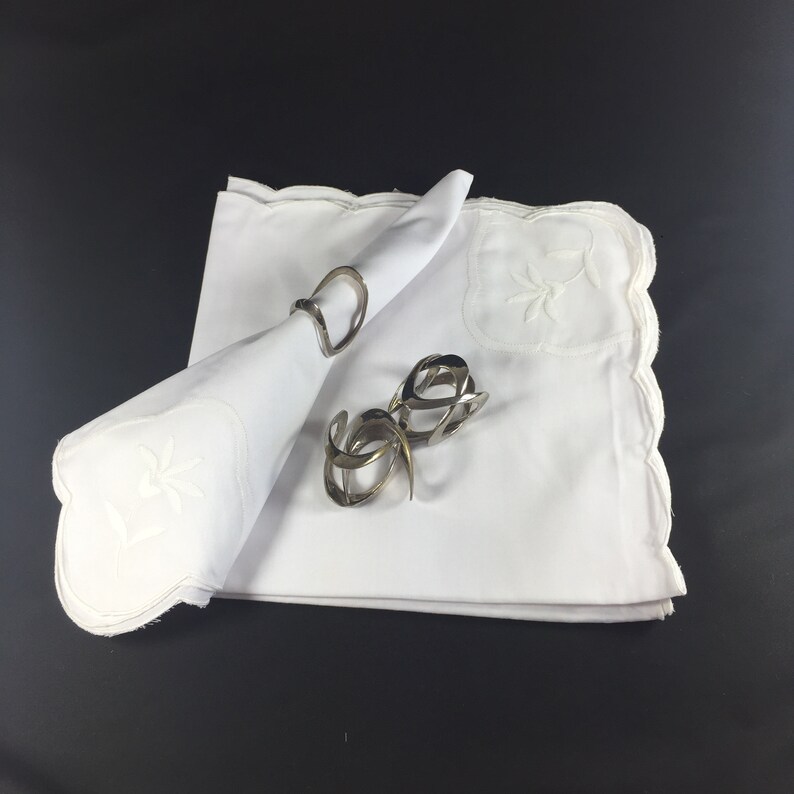 May include: Two silver napkin rings on a white linen napkin with embroidered floral designs. The napkin is folded into a triangle shape.