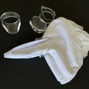 May include: Clear acrylic napkin rings with a white cloth napkin. The napkin has a decorative lace edge. The napkin rings are a modern design, and the napkin is folded with a point.