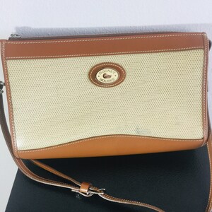May include: A tan and brown Dooney & Bourke crossbody bag with a woven fabric front and a leather flap. The bag has a gold metal logo with the text "Dooney & Bourke".