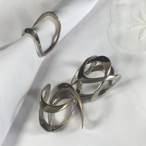 May include: Two silver napkin rings with a modern, abstract design. The rings are made of metal and have a smooth, polished finish.