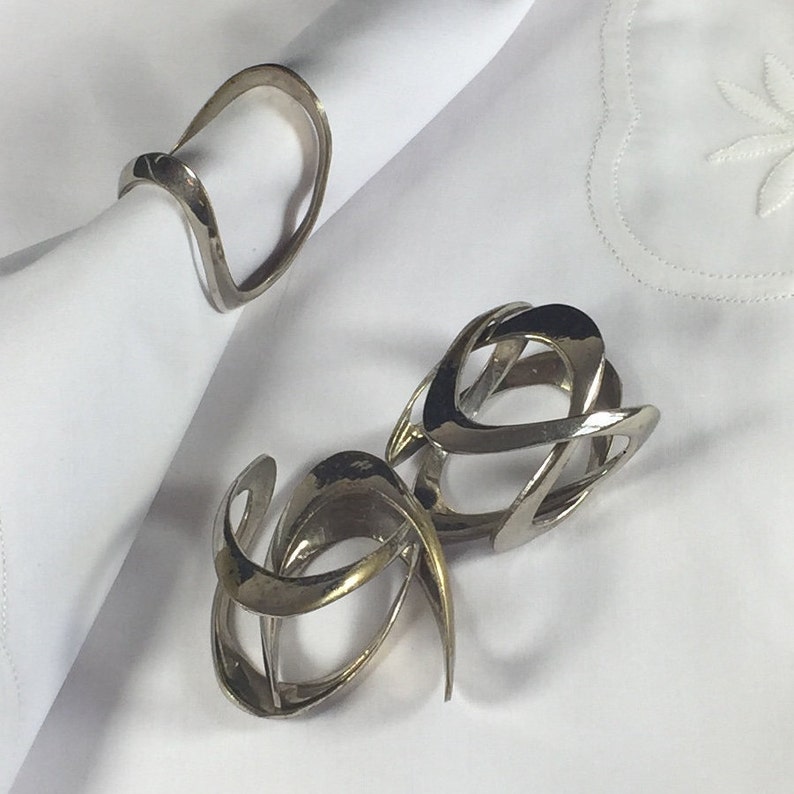 May include: Two silver napkin rings with a modern, abstract design. The rings are made of metal and have a smooth, polished finish.