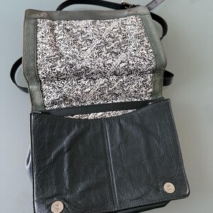 May include: A black leather crossbody bag with a fold-over flap featuring two silver magnetic snap closures. The interior lining is white with a repeating black graphic pattern and the text REBECCAMINKOFF. It includes a thin black shoulder strap.