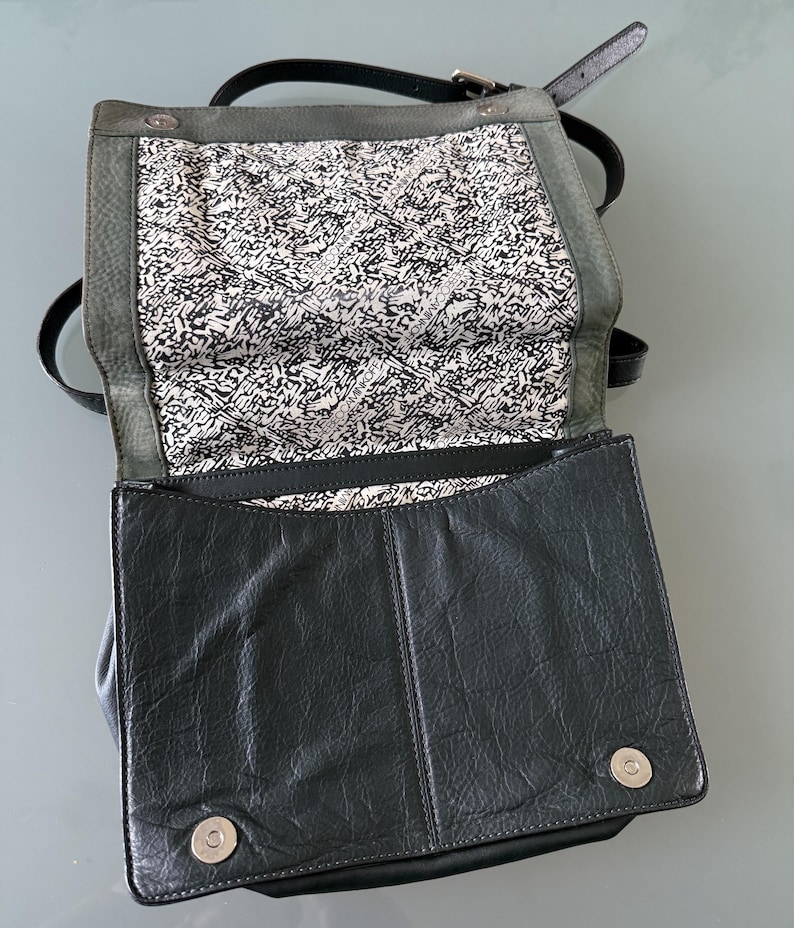 May include: A black leather crossbody bag with a fold-over flap featuring two silver magnetic snap closures. The interior lining is white with a repeating black graphic pattern and the text REBECCAMINKOFF. It includes a thin black shoulder strap.