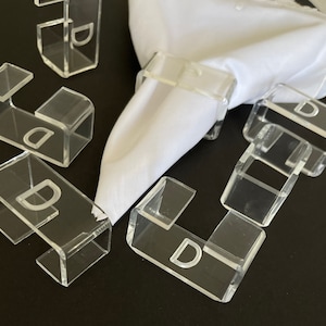 May include: Set of clear acrylic napkin rings with the letter "D" engraved on each ring. The rings are designed to hold napkins in place.