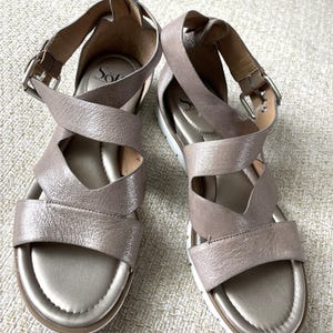 May include: A pair of women's sandals with a metallic taupe finish. The sandals feature crisscross straps, adjustable ankle straps with buckles, and white soles. The brand name "So" is visible on the footbed.