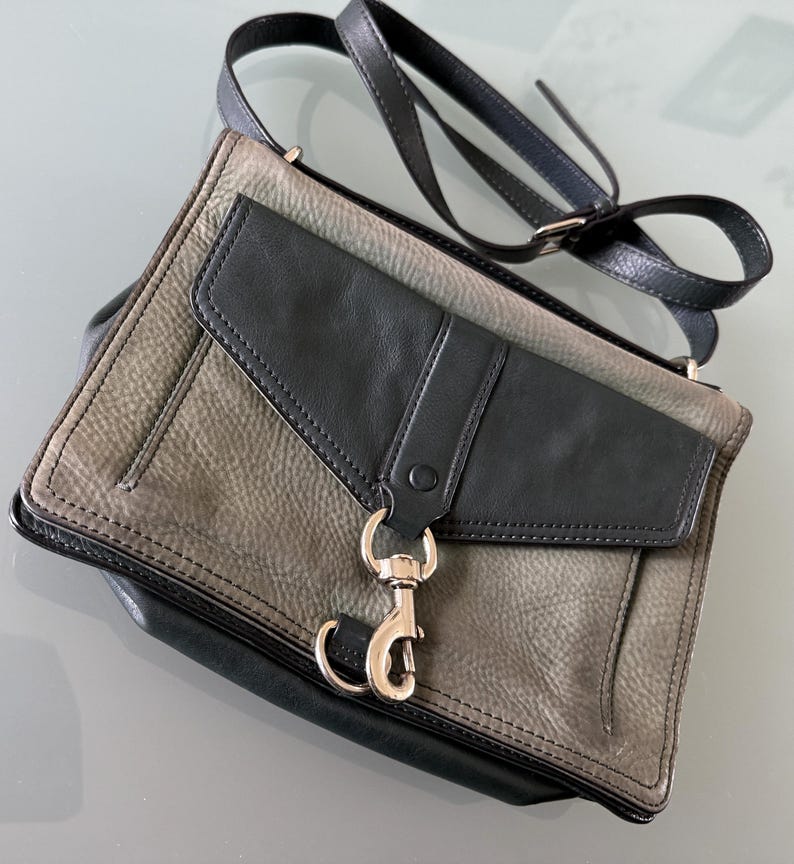 May include: A two-tone crossbody bag featuring a textured grey leather body with a smooth black leather flap. The bag includes a silver-toned metal lobster clasp closure on the front, a matching black leather shoulder strap, and black leather trim.