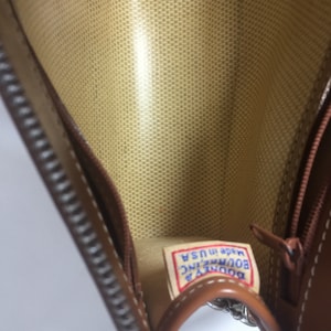 May include: The inside of a brown leather purse with a zipper closure. The lining is a beige fabric with a woven pattern. A blue and white label reads "Dooney & Bourke INC. Made in USA."