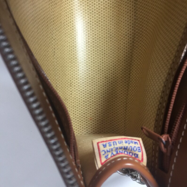 May include: The inside of a brown leather purse with a zipper closure. The lining is a beige fabric with a woven pattern. A blue and white label reads "Dooney & Bourke INC. Made in USA."
