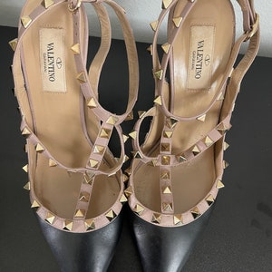 May include: A pair of black and nude leather high-heeled shoes with gold studs. The shoes have a pointed toe and an ankle strap. The brand name "VALENTINO GARAVANI" is printed on the insole.