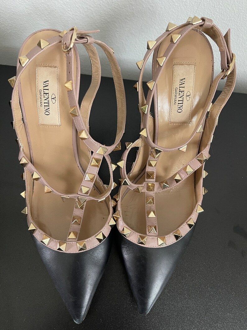 May include: A pair of black and nude leather high-heeled shoes with gold studs. The shoes have a pointed toe and an ankle strap. The brand name "VALENTINO GARAVANI" is printed on the insole.