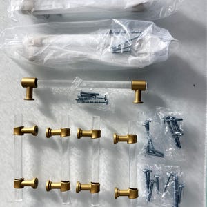 May include: A collection of clear acrylic and gold-toned cabinet hardware, including handles and pulls. The hardware is packaged in clear plastic, with screws and mounting hardware visible. The handles and pulls are in various sizes.