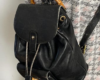 May include: Black leather backpack with a bamboo handle and drawstring closure. The bag has a flap with a gold buckle and a small pocket on the front.