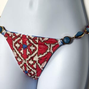 May include: A red and white patterned bikini bottom with blue accents. The bikini bottom is adorned with a chain of blue gemstone beads and gold-colored metal links. The fabric has a geometric design.