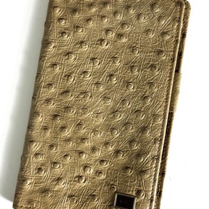 May include: Tan leather wallet with a textured, ostrich-like pattern. The wallet has a rectangular shape with rounded corners and a small, rectangular metal accent. The wallet is closed and appears to be new.
