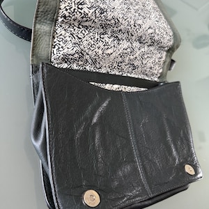 May include: A black leather handbag with a textured surface, featuring a fold-over flap secured by two silver-toned magnetic snap buttons. The interior lining is white with a black abstract graphic print and the text Rebecca Minkoff.
