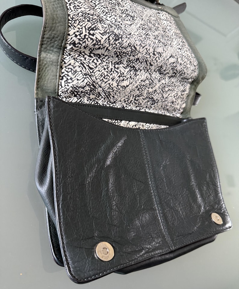 May include: A black leather handbag with a textured surface, featuring a fold-over flap secured by two silver-toned magnetic snap buttons. The interior lining is white with a black abstract graphic print and the text Rebecca Minkoff.