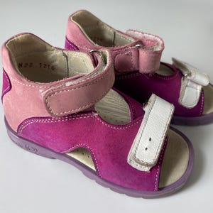 May include: A pair of pink and white sandals with velcro straps. The sandals have the size "N 25 1216" written on the inside.