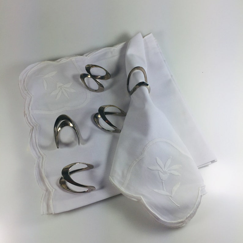 May include: Set of six silver napkin rings with a modern design. The rings are placed on a white linen napkin with embroidered floral details.