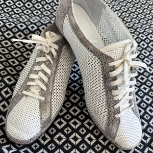 May include: A pair of white and gray sneakers with white laces. The sneakers have a perforated design on the sides and toe, with gray accents around the edges. The shoes are displayed on a black and white patterned surface.