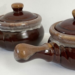 May include: Two brown ceramic soup tureens with lids and handles. The tureens have a white glaze around the top edge and a brown glaze on the rest of the body. The handles are brown with a white glaze on the ends.