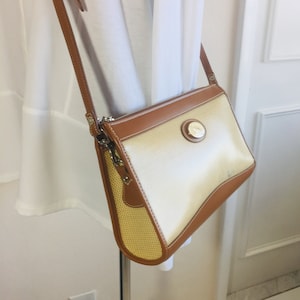 May include: A tan and brown crossbody bag with a woven fabric front and a brown leather trim. The bag has a zipper closure and a circular logo on the front.