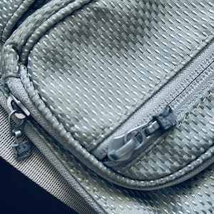 K&ouml;nnte beinhalten: Nahaufnahme eines grauen Rei&szlig;verschlusses an einer silbernen, strukturierten Stofftasche. Der Rei&szlig;verschluss hat einen silbernen Schieber und einen schwarzen Rei&szlig;verschluss-Kopf.