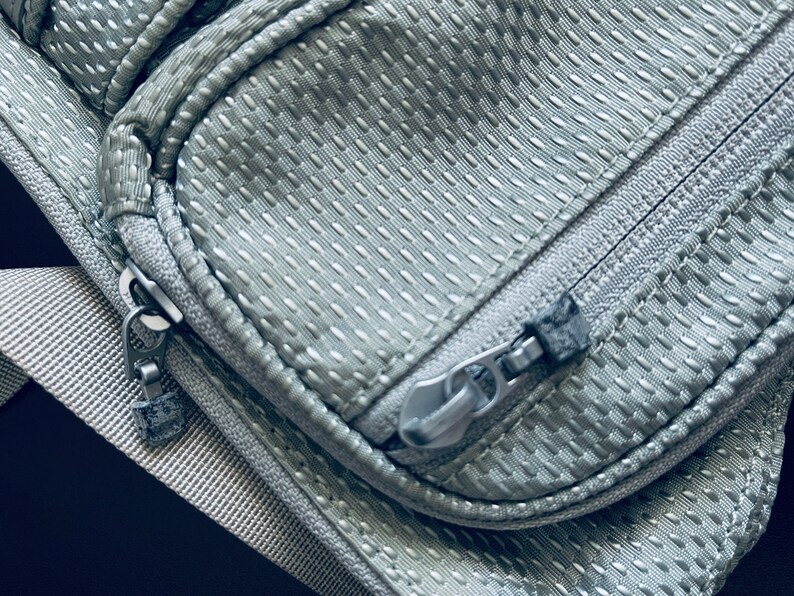 K&ouml;nnte beinhalten: Nahaufnahme eines grauen Rei&szlig;verschlusses an einer silbernen, strukturierten Stofftasche. Der Rei&szlig;verschluss hat einen silbernen Schieber und einen schwarzen Rei&szlig;verschluss-Kopf.