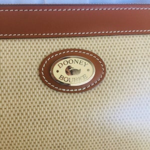 May include: A close-up of a Dooney & Bourke brand logo on a beige fabric bag. The logo is a gold oval with the words "Dooney & Bourke" and a small duck image.