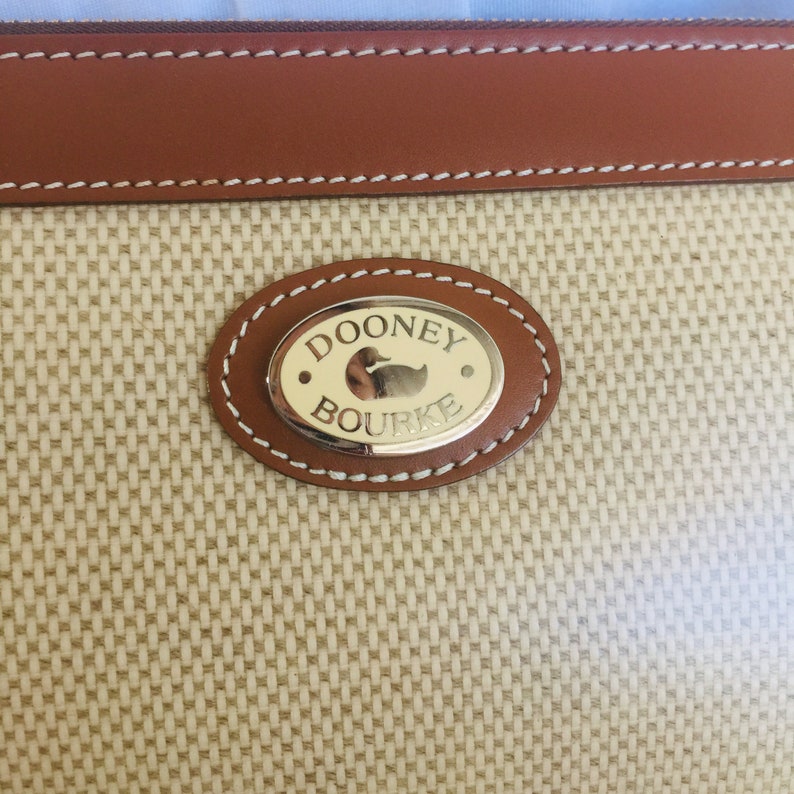 May include: A close-up of a Dooney & Bourke brand logo on a beige fabric bag. The logo is a gold oval with the words "Dooney & Bourke" and a small duck image.