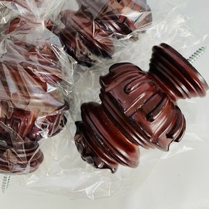 May include: Three dark brown wooden finials with intricate carvings and a threaded metal screw. The finials are packaged in clear plastic.